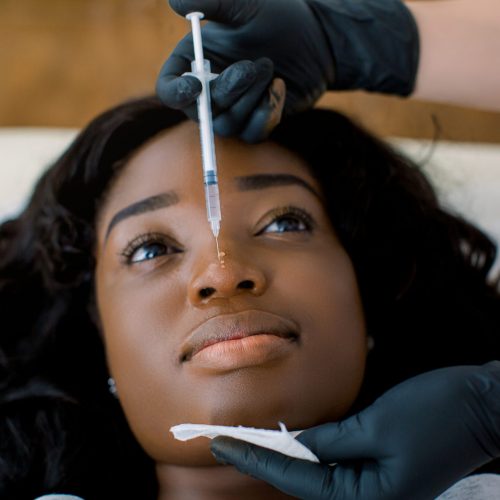 Close up view of hands of female doctor cosmetologist making rejuvenating therapy, facial injections procedure for tightening and smoothing face skin of a beautiful, young African woman in clinic.